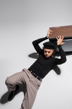 Siyah balıkçı yakalı ve pantolonlu Afro-Amerikan adam gri arka planda yalan söylüyor ve eski bir televizyona dokunuyor.