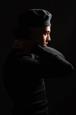 side view of african american man in pullover and beret posing with hands behind neck isolated on black clipart