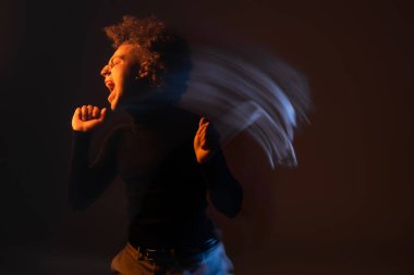 long exposure of angry and stressed african american man with bipolar disorder screaming on black background with orange and blue light clipart