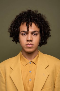 Esmer, Afro-Amerikalı, sarı gömlekli ve ceketli bir adam, gri renkli kameraya bakıyor.