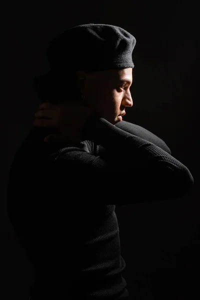side view of african american man in pullover and beret posing with hands behind neck isolated on black