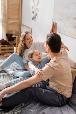 cheerful kid looking at father giving high five to happy mother while sitting on bed 
