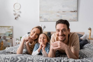 portrait of joyful parents and kid looking at camera while resting in bedroom 