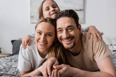 portrait of happy child hugging cheerful parents looking at camera while resting in bedroom 
