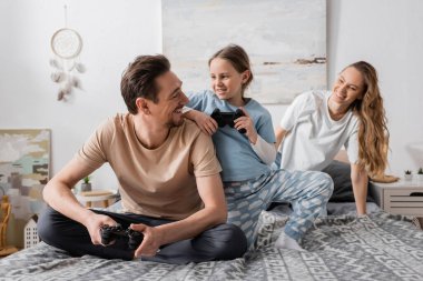 KYIV, UKRAINE - NOVEMBER 28, 2022: cheerful father and child playing video game near wife in bedroom 