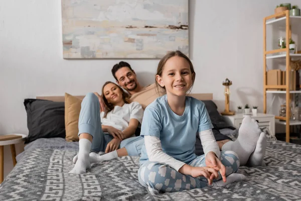 stock image cheerful girl sitting on bed near blurred parents resting on background 