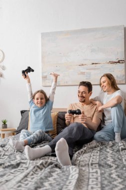 KYIV, UKRAINE - NOVEMBER 28, 2022: cheerful woman leaning on shoulder of husband playing video game with excited daughter 