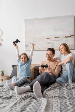 KYIV, UKRAINE - NOVEMBER 28, 2022: cheerful woman leaning on shoulder of husband playing video game with excited daughter 