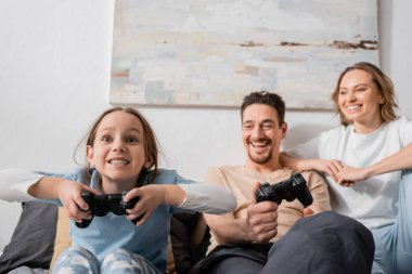 KYIV, UKRAINE - NOVEMBER 28, 2022: happy parents looking at excited daughter playing video game 