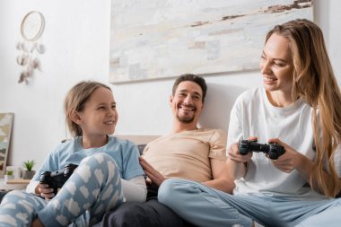 KYIV, UKRAINE - NOVEMBER 28, 2022: cheerful woman and child playing video game near husband in bedroom 