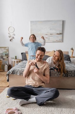 KYIV, UKRAINE - NOVEMBER 28, 2022: happy parents playing video game near excited daughter in bedroom 