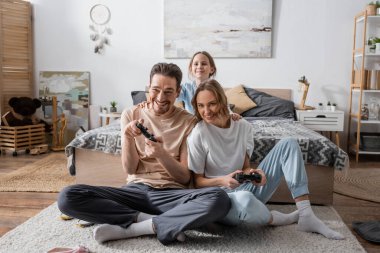 KYIV, UKRAINE - NOVEMBER 28, 2022: cheerful parents playing video game near smiling daughter in bedroom 