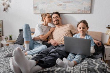 cheerful parents looking at happy kid watching movie on laptop in bedroom 