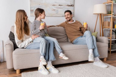 full length of cheerful daughter sitting on laps of mother and holding glass of orange juice near positive father 