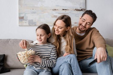 smiling kid looking at bowl while happy father taking popcorn near cheerful mother 