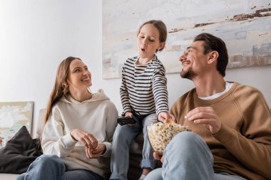 kid holding remote controller and reaching popcorn near happy parents 