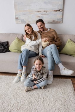 amazed kid clicking channels near happy parents eating popcorn at home  