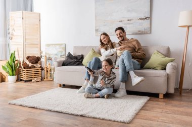 full length of cheerful kid clicking channels near happy parents sitting with popcorn at home  