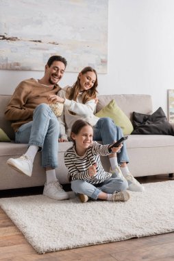 full length of positive kid clicking channels near happy parents sitting with popcorn at home  