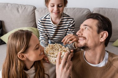 happy man reaching popcorn near wife and cheerful daughter on sofa 