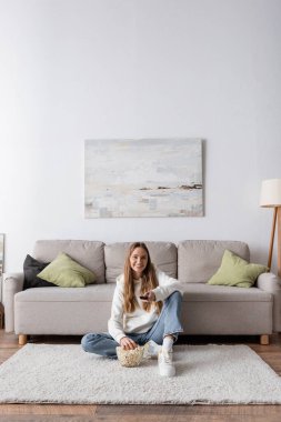 full length of cheerful woman holding remote controller while watching movie in living room  clipart