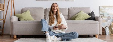 full length of joyful woman holding remote controller while watching movie in living room, banner 