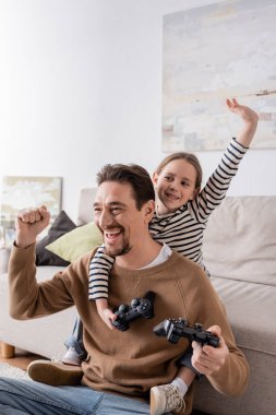 KYIV, UKRAINE - NOVEMBER 28, 2022: cheerful father and daughter holding joysticks and rejoicing after video game 