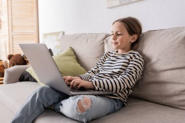 preteen girl in casual clothes using laptop while studying online and sitting on couch 