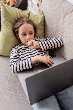 high angle view of focused preteen girl in casual clothes using laptop while lying on couch 