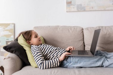 side view of cheerful preteen girl in casual clothes using laptop while lying on couch 