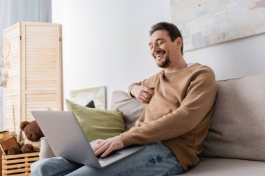 cheerful man using laptop while sitting on sofa in living room 