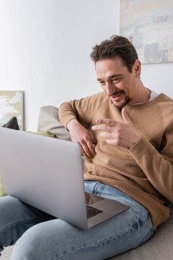 bearded man using laptop while having video call and sitting on sofa in living room 