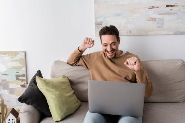 positive man watching championship on laptop while cheering and sitting on sofa in living room 