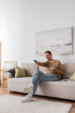 full length of cheerful man holding remote controller and cup of coffee in living room 