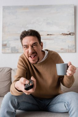 tensed man holding remote controller and cup of coffee in living room 