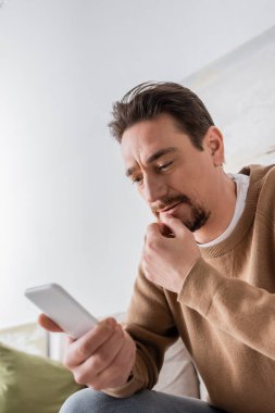 low angle view of bearded man in beige jumper messaging on smartphone at home 