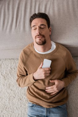 top view of bearded man with closed eyes holding smartphone and leaning on sofa 