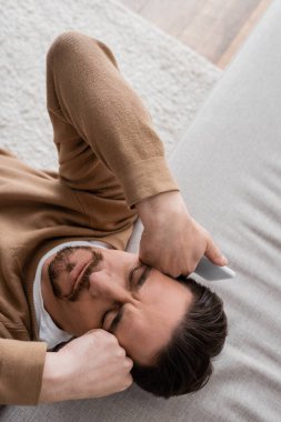 top view of tired man in beige jumper holding mobile phone and leaning on sofa 