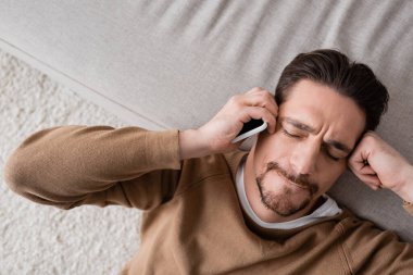 top view of exhausted man in beige jumper holding mobile phone and leaning on sofa 