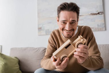 joyful man smiling while looking at camera and holding wooden car toy in living room 