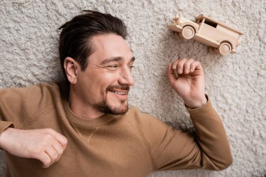 top view of bearded and cheerful man lying on carpet near wooden toy car in living room 