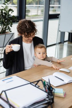 businesswoman sitting with little daughter and coffee cup near documents and blank notebook in office clipart