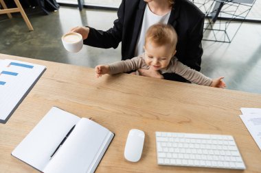 high angle view of cheerful baby near mother with coffee cup and computer mouse with keyboard and blank notebook on office desk