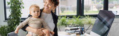 smiling businesswoman holding toddler daughter near computer monitor with blank screen in office, banner clipart