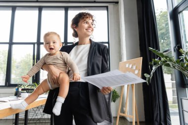 happy businesswoman in black suit looking away while standing with baby and documents in office clipart