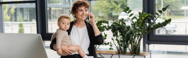 smiling businesswoman with little daughter talking on smartphone near computer monitor and green plants in office, banner