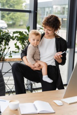 smiling businesswoman with baby and mobile phone near blurred notebook and coffee cup on work desk