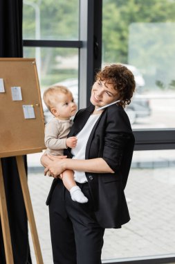 positive businesswoman in black suit holding little daughter and talking on smartphone in office clipart