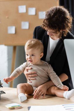 cheerful baby in romper sitting on office desk near smiling mother and sticky notes clipart