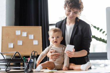 little baby in romper sitting on desk near smiling mom in black blazer using mobile phone in office